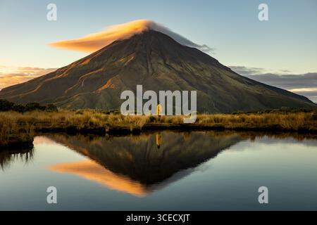Vista laterale di una persona irriconoscibile in piedi accanto a un lago riflettente, guardando il maestoso Monte Taranaki durante l'alba in nuova Zelanda. A Serene e tr Foto Stock
