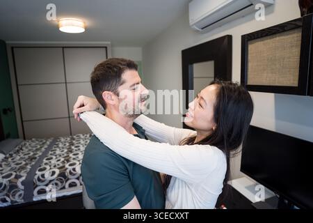 Una coppia diversificata condivide un momento tenero a casa, abbracciando un ambiente interno accogliente. Il decor moderno e l'atmosfera calda evidenziano il loro legame Foto Stock