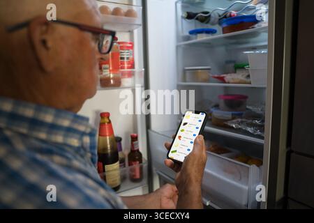 Vista laterale di un uomo anziano irriconoscibile che utilizza uno smartphone per acquistare generi alimentari online, davanti a un frigorifero aperto per controllare gli articoli da ordinare. Foto Stock