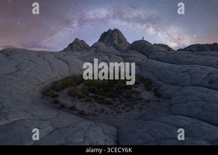 Un uomo solitario si trova sotto la vastissima via Lattea nel terreno ultraterreno di White Pocket, Arizona. Il cielo notturno illumina il paesaggio desertico Foto Stock