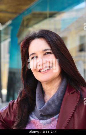 Una donna sicura di sé con lunghi capelli scuri sorride calorosamente, vestita con un'elegante giacca rossa e sciarpa grigia. È posizionata su uno sfondo urbano moderno Foto Stock