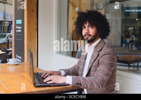 Venditore di assicurazioni miste con capelli afro che utilizza un computer portatile a un tavolo di legno in un bar. Con un blazer a motivi geometrici, concentrato e professionale in una Foto Stock