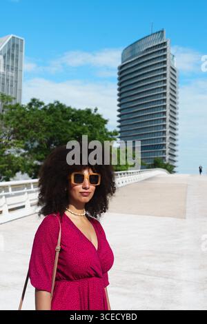 Giovane donna araba con capelli afro che indossa un abito rosso e occhiali da sole, all'aperto con edifici moderni sullo sfondo in una giornata di sole. Foto Stock