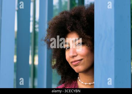 Una giovane donna araba con i capelli ricci sorride guardando con sicurezza la macchina fotografica, incorniciata da colonne blu. La sua espressione trasuda calore e fascino, catturando un m Foto Stock