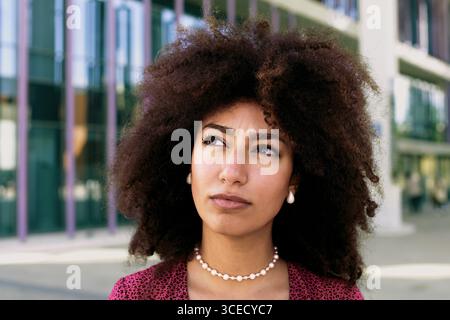 La giovane donna araba con i capelli afro ricci si trova all'aperto, sembra premurosa. Indossa una collana di perle e orecchini, con un'architettura moderna Foto Stock