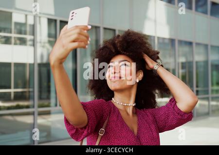 Una giovane donna araba sorride con fiducia mentre scatta un selfie utilizzando il suo smartphone all'esterno di un edificio moderno, evidenziando la sua espressione gioiosa e. Foto Stock