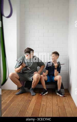 Un legame padre e figlio mentre si fa esercizio con i manubri a casa, seduti su una panchina in una stanza luminosa. L'interazione enfatizza fitness, unità e hea Foto Stock