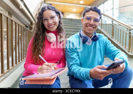 Una giovane e allegra donna della generazione Z con cuffie e un tablet e un uomo con cuffie che reggono uno smartphone, siediti sulle scale, incarnando la moderna vita digitale Foto Stock