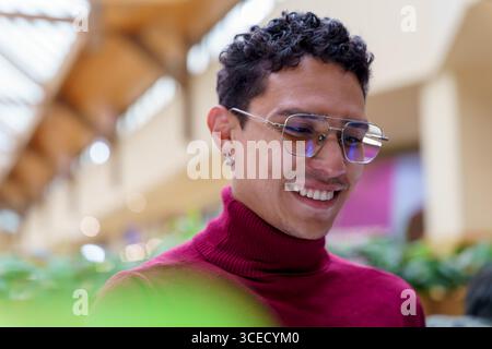 Un giovane felice della Gen Z sorride brillantemente in uno spazio pubblico al coperto, mostrando uno stile di vita allegro e moderno con un ambiente vibrante nella ba Foto Stock