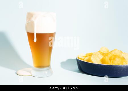 Un bicchiere alto di birra con schiuma che scorre sopra, posto accanto a una ciotola di patatine fritte. La scenografia è impostata su uno sfondo azzurro, creando un rinfrescante Foto Stock