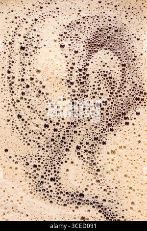 Immagine ravvicinata che cattura la trama intricata e frizzante della schiuma di caffè su una superficie color beige cremosa. Lo schema formato dalle piccole bolle crea una m. Foto Stock