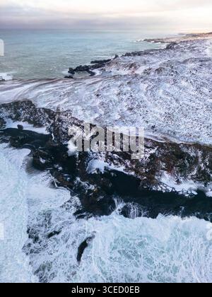 Vista aerea dell'aspra costa islandese in inverno, che mostra il drammatico contrasto tra le rocce vulcaniche spolverate di neve e il blu ghiacciato dell'Atla Foto Stock