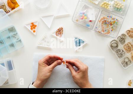 Vista dall'alto delle mani irriconoscibili ritagliate di una donna che crea gioielli nel suo studio. Circondata da perline e strumenti, crea con attenzione e abilità Foto Stock