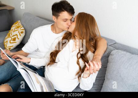 Una coppia incinta abbraccia tenerezza mentre condivide un bacio amorevole su un comodo divano, evidenziando il calore e l'intimità dei momenti familiari e futu Foto Stock