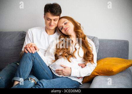 Una giovane coppia abbraccia tenerezza sul divano, irradiando gioia e anticipazione mentre non vedono l'ora di accogliere il loro primo bambino in famiglia, symb Foto Stock