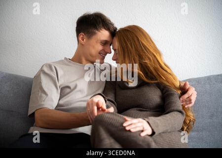 Una giovane coppia che aspetta un bambino condivide un momento tenero sul divano, abbracciando felicità e attesa. Le loro espressioni gioiose riflettono l'amore e l'ex Foto Stock