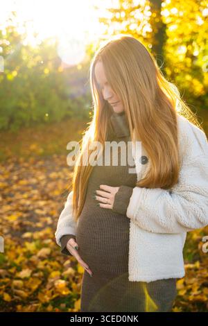 Una bella donna incinta con i capelli rossi si trova pacificamente in mezzo a un paesaggio autunnale illuminato dal sole, incarnando calore e serenità mentre le foglie dorate circondano la h Foto Stock