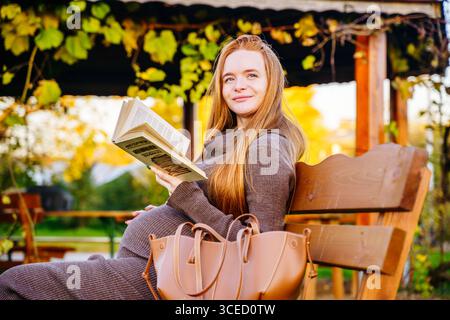 Una bella donna incinta con i capelli rossi siede su una panchina del parco, godendo un libro. La scena cattura l'essenza di un autunno tranquillo, evidenziato da vibr Foto Stock