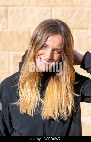 Una giovane donna con i capelli lunghi sorride brillantemente alla luce del sole, indossando una giacca nera. La parete in mattoni beige sullo sfondo aggiunge calore al cheerf Foto Stock