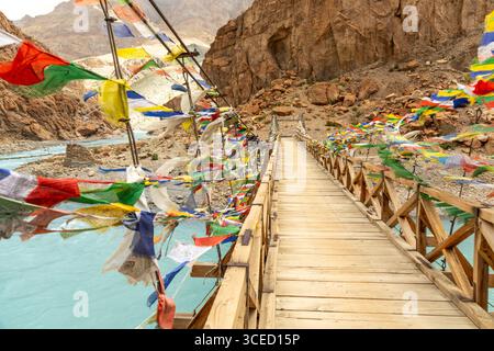 Un ponte sospeso adornato da vibranti bandiere di preghiera si estende su un fiume turchese nel terreno accidentato del Ladakh, che conduce verso la remota Phug Foto Stock