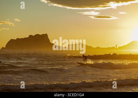 Un surfista si staglia sullo sfondo di uno splendido tramonto sul Mar Mediterraneo al largo della Costa Blanca. L'acqua tranquilla e il cielo vibrante crea Foto Stock