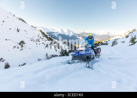 Una motoslitta naviga attraverso paesaggi invernali incontaminati tra le montagne, circondati da alberi e vette innevate, catturando l'essenza di adve Foto Stock