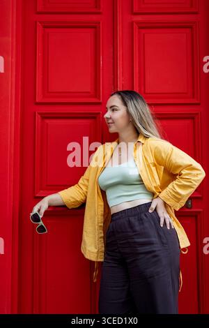 Una donna alla moda con una camicia gialla vibrante si erge con sicurezza contro una porta rossa brillante su una strada della città, incarnando l'essenza dell'esplorazione urbana Foto Stock