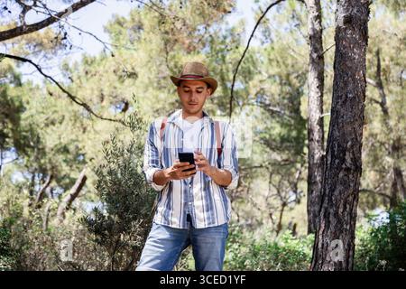 Un giovane uomo, vestito in modo informale con un cappello, si trova in una foresta lussureggiante utilizzando il suo smartphone. Trascorre una giornata tranquilla nella natura, esplorando e catturando Foto Stock