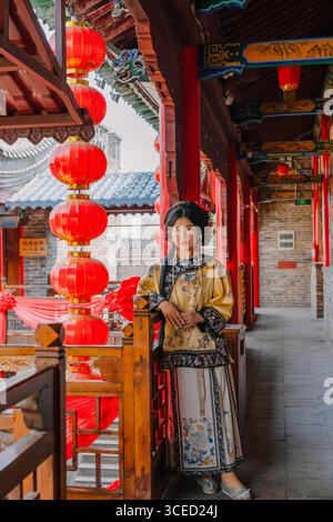 Donna in tradizionale abbigliamento della dinastia Qing si erge graziosamente in un corridoio storico a Pingyao, in Cina, circondato da lanterne rosse e intricato architec Foto Stock