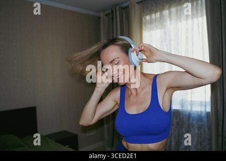 Una donna sta ballando gioiosamente a casa, indossando cuffie bianche. I suoi occhi sono chiusi mentre si muove verso la musica, sentendosi rilassata e spensierata in un'occasione Foto Stock