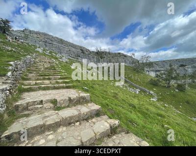 I gradini di pietra di Malham salgono su una collina erbosa, che conduce verso una montagna rocciosa sotto un cielo blu nuvoloso. Foto Stock