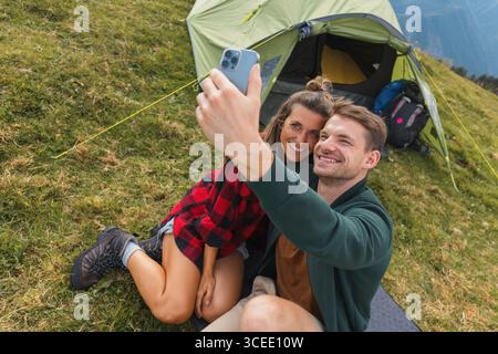 Coppia romantica in campeggio che cattura i momenti all'aperto con uno smartphone, seduto accanto a una tenda in un paesaggio di montagna panoramico. Foto Stock