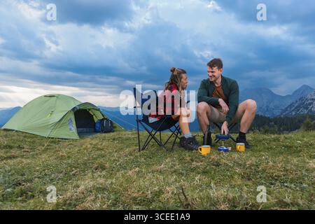 Giovani coppie che si godono una mattinata in campeggio spensierata, si divertono davanti alla loro tenda, preparando pancake su un bruciatore a gas. Foto Stock