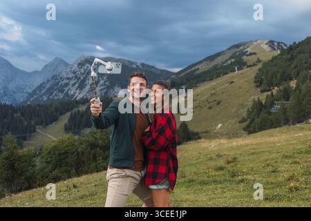 Una giovane coppia che scatta selfie, saluta e manda baci mentre effettua una videochiamata sulla cima di una montagna con un paesaggio panoramico sullo sfondo. Foto Stock