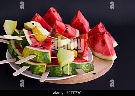 Anguria fresca e matura e melone tagliati su piatto di legno con semi di anguria, bastoncini di legno su sfondo nero Foto Stock