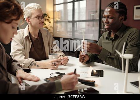 Gruppo di giovani uomini e donne diversi seduti al tavolo in ufficio, discutendo la progettazione di turbine eoliche Foto Stock