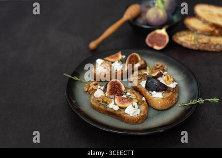 Vista frontale orizzontale di tre toast di formaggio di fico e capra su piatto verde con timo, noci, cucchiaio di miele, saporito mix dolce, sfondo scuro Foto Stock