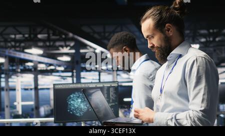 Tecnico che tiene un notebook in movimento sulla piattaforma server farm, automatizzando l'apprendimento automatico dell'intelligenza artificiale. Programmatore di data center che utilizza un notebook, esegue la manutenzione utilizzando l'intelligenza artificiale Foto Stock