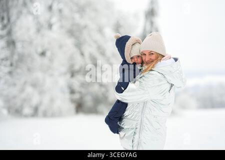 Madre che tiene il bambino in un intimo abbigliamento invernale in un paesaggio sereno e innevato. Entrambi sono vestiti con calore, esprimendo gioia e calore all'aperto. Perfetto Foto Stock