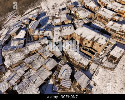 La vista aerea dei tetti polverati di neve degli edifici raggruppati crea un mosaico strutturato a contrasto con il paesaggio bianco scuro, Ostana, Piemonte, Italia. Foto Stock