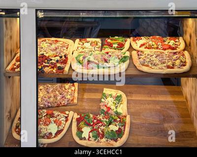 Esposizione di pizze rettangolari con vari condimenti all'interno di una vetrina di vetro al deli. Concetto di esposizione di pizza, fast food cittadino, opzione pranzo veloce Foto Stock