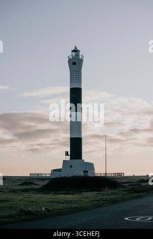 Un alto faro si erge da solo su una collina erbosa. Il cielo è limpido e il sole tramonta, proiettando un caldo bagliore sulla scena. Il faro è un simbolo Foto Stock