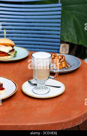 Tazza di cappuccino frizzante servito su un rustico tavolo da caffè all'aperto, accompagnato da piatti con sandwich per la colazione, pasticcini e dessert. Accogliente Foto Stock