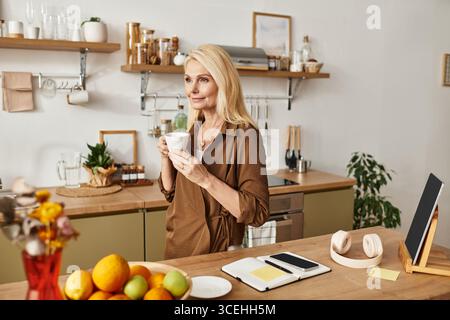 Una donna anziana con lunghi capelli biondi sorseggia il tè mentre ammira la sua elegante cucina piena di frutta. Foto Stock