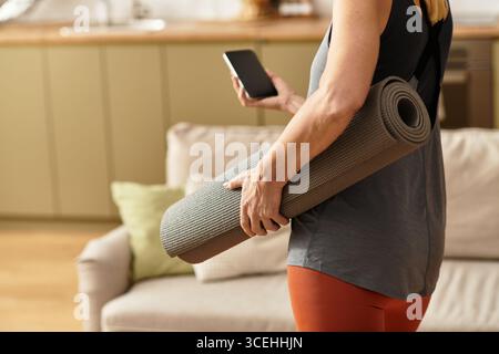 Una donna anziana con un tappetino per lo yoga e uno smartphone si rilassa nel suo spazio di vita consapevole. Foto Stock