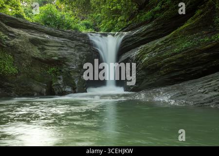 Vista di una vigorosa cascata che si getta su rocce aspre in una tranquilla piscina, circondata da lussureggiante vegetazione, creando un vibrante contrasto di texture, Mirsha Foto Stock