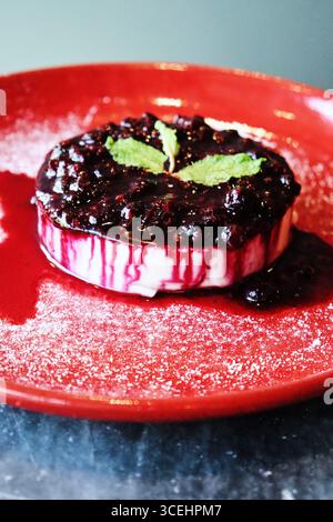 Classico dessert italiano alla panna cotta servito su un piatto rosso vivace, condito con una ricca salsa di frutti di bosco e frutta fresca. Foto Stock