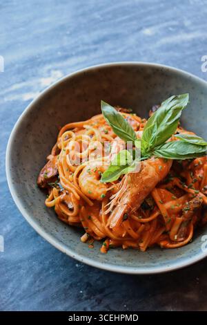 Primo piano di una deliziosa pasta a base di pesce con gamberi in una ricca salsa di pomodoro, guarnita con foglie di basilico fresco ed erbe aromatiche, servita in una rustica ciotola di ceramica Foto Stock