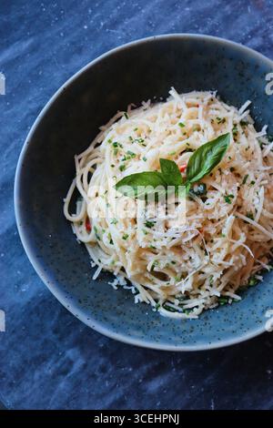 Primo piano di classici spaghetti italiani conditi con parmigiano grattugiato, foglie di basilico fresco ed erbe aromatiche, serviti in una rustica ciotola di ceramica blu. Foto Stock