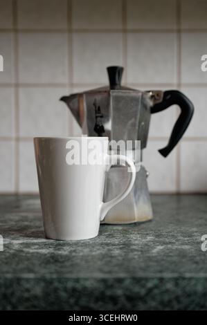 Tazza bianca semplice di fronte ad una piccola macchina per il caffè Bialetti Moka Express su un piano cucina a motivi verdi-grigi, con piastrelle bianche sullo sfondo. Foto Stock